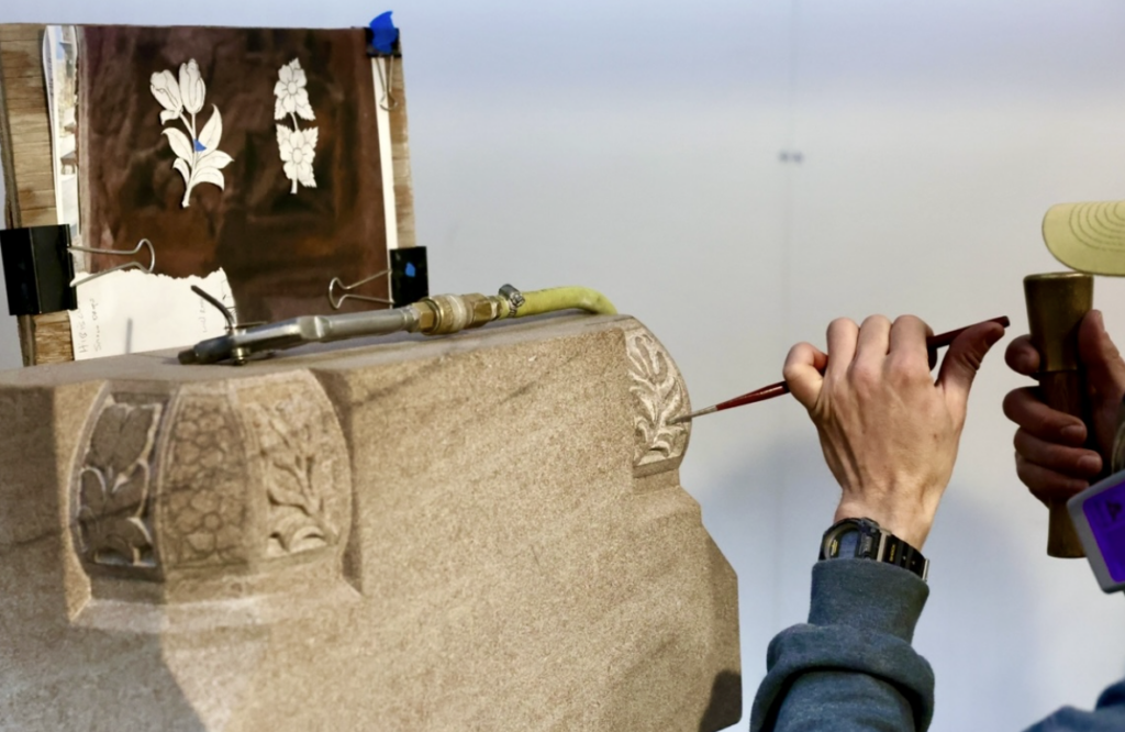 A person is using a chisel and hammer to carve intricate floral designs into a natural stone surface. A reference sketch of flowers is clipped nearby, adding to the timeless beauty of this focused craftsmanship in stone carving.